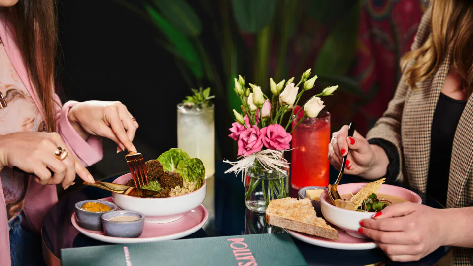 Guests dining at an elegant restaurant table, enjoying fresh salads, bread, and drinks, with vibrant flowers as decor.