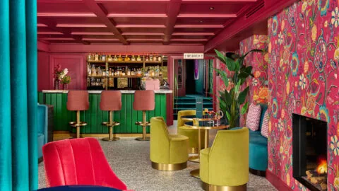 Vibrant hotel lounge with colourful seating, floral wallpaper, cosy fireplace, and fully stocked bar, inviting atmosphere.