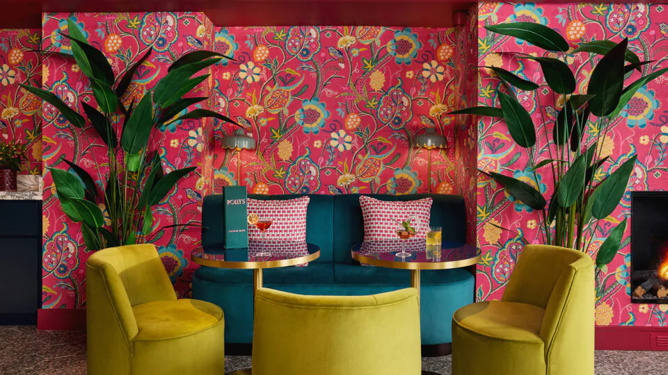 Vibrant hotel lounge with pink floral wallpaper, green chairs, and a warm fireplace, offering a cosy, welcoming ambience.