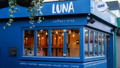 Cosy café exterior at dusk, featuring blue walls, warm lights, offering coffee, wine, and pastries for a relaxing experience.