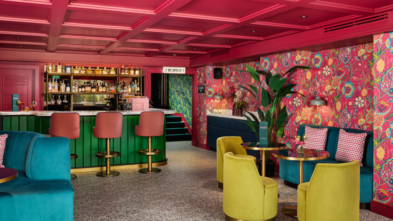 A vibrant bar lounge features colorful patterned wallpaper, green and blue seating, gold accents, and a well-stocked bar with green paneling and pink overhead details.